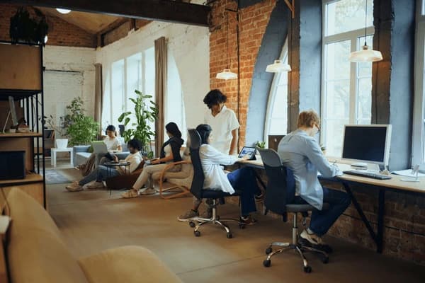Team collaborating and working on laptops in a creative office space with exposed brick, wooden beams, and large windows.