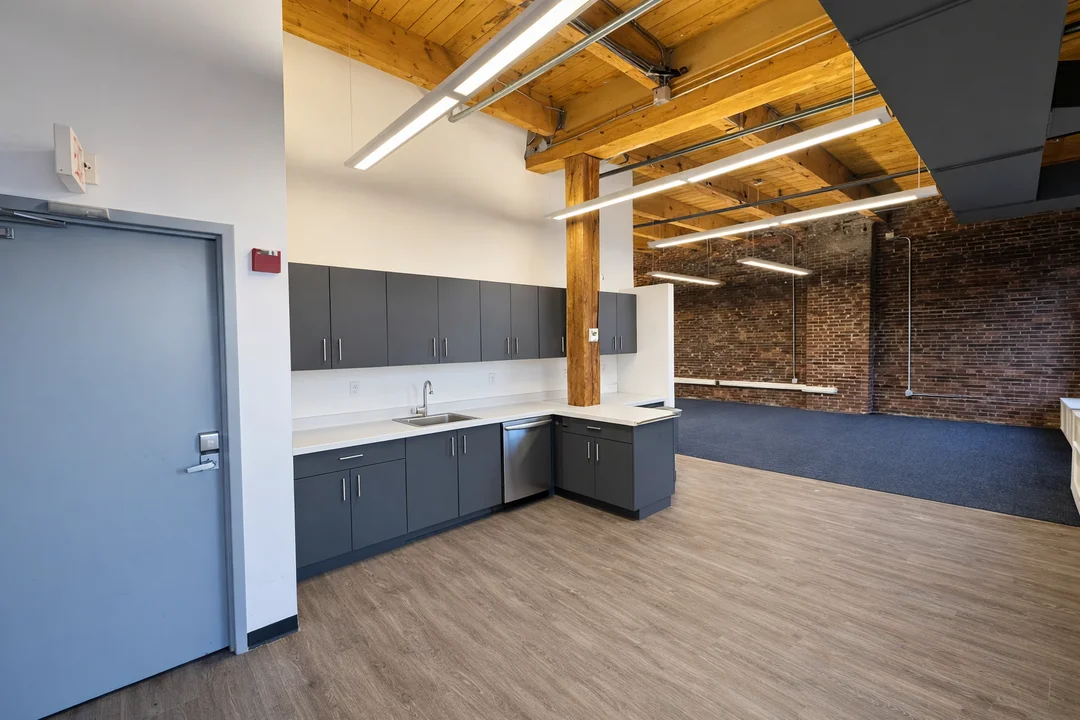 Renovated loft office interior featuring exposed brick, timber beam ceilings, modern lighting, and a built-in kitchenette.
