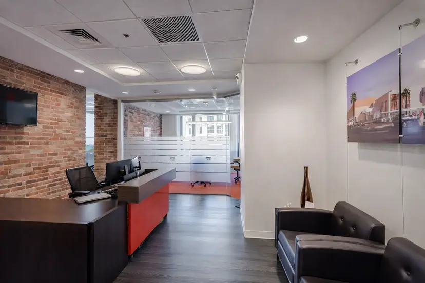Modern office lobby featuring a reception desk, exposed brick accent walls, glass conference room, and black leather seating.