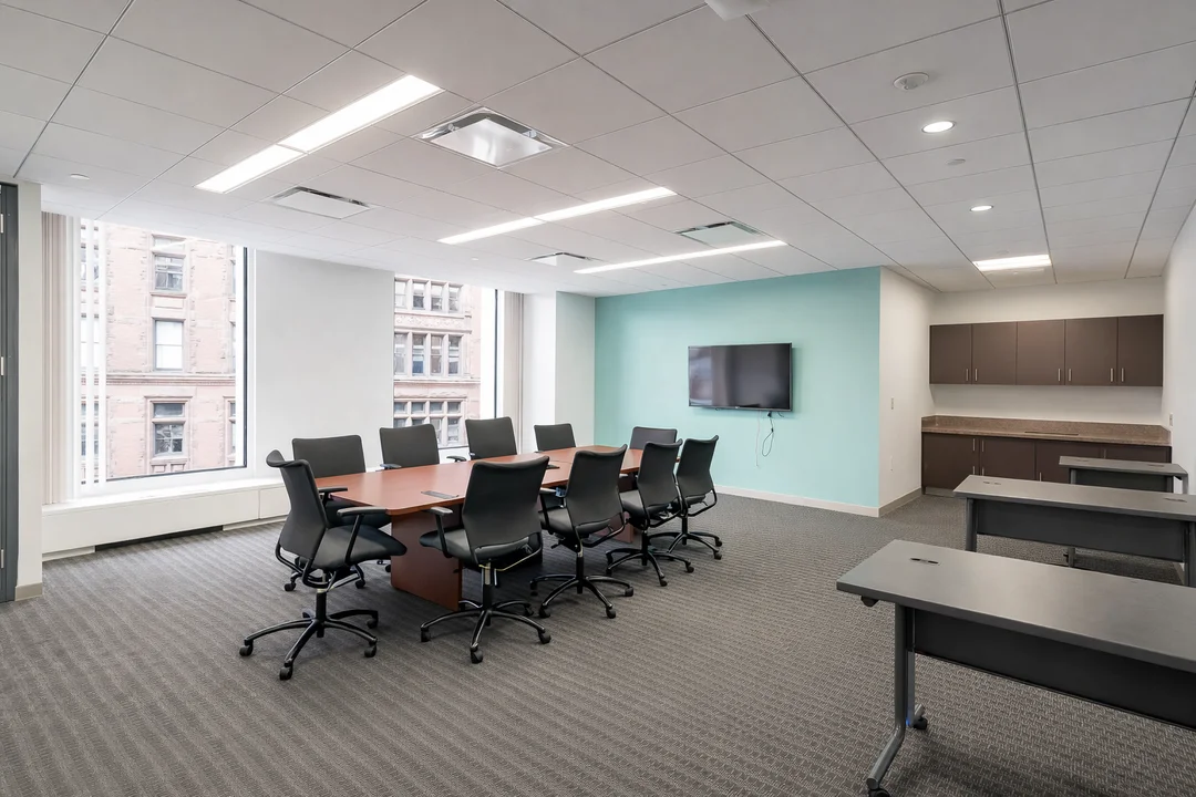 Bright office conference room featuring a wood meeting table, rolling chairs, large windows, and a wall-mounted flat-screen display.